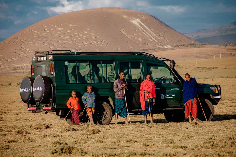 Maasai men and boys standing with the Oloitiko safari vehicle below a volcanic cone.